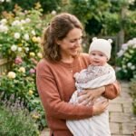 Una mamá con su bebé en un jardín lleno de flores y plantas de hoja verde.
