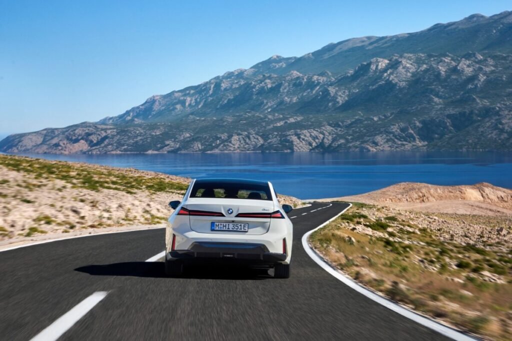 Así es el nuevo BMW i3 50 xDrive: la berlina eléctrica de los 906 km de autonomía que ya puedes reservar Imagen que muestra el nuevo BMW i3 de color blanco, circulando por una carretera. Vista parte trasera del vehículo.