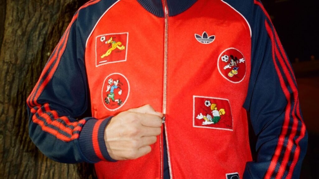 El calzado híbrido de adidas con Brain Dead que reimagina el fútbol de los 90 para la calle Imagen que muestra una sudadera roja de adidas llena de parches de disney de la nueva colaboración de 2026
