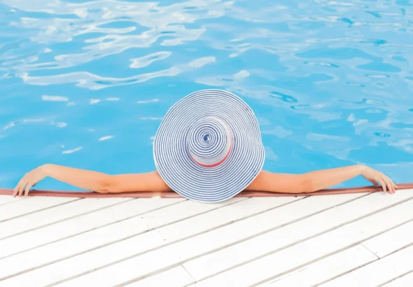 Flamingueo pone de moda la playa con sus accesorios de color rosa. Toallas, flotadores, sombrillas... la imagen muestra a una chica en una piscina con un gorro preparando sus complementos de Flamingueo